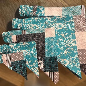 Puede incluir: Una colección de cinco banderines de tela de diferentes tamaños, dispuestos en una exhibición en capas. Cada banderín presenta un diseño de patchwork con patrones turquesa, blanco, gris y negro, incluidos motivos florales y geométricos. Los banderines tienen tela fruncida en la parte superior.