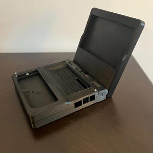 Puede incluir: Un dispositivo plegable negro impreso en 3D que se asemeja a un ordenador portátil en miniatura. Cuenta con una base rectangular con un compartimento empotrado y una pantalla abatible. El exterior tiene un acabado texturizado con rejillas laterales.