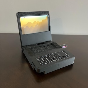 Puede incluir: Un dispositivo informático portátil negro con un marco texturizado impreso en 3D, que cuenta con una pantalla integrada que muestra un fondo de escritorio de paisaje, un teclado QWERTY compacto y un pequeño panel táctil lateral.
