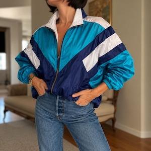 Può includere: Una giacca a vento con zip caratterizzata da un design color-block in turchese brillante, blu navy intenso e bianco. La giacca presenta un colletto alto, maniche lunghe con polsini elastici e una chiusura frontale con zip, abbinata a jeans blu classici.