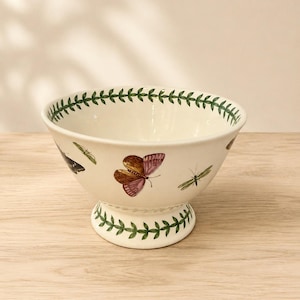 May include: A cream-colored ceramic footed bowl featuring a decorative green leaf vine pattern along the rim and base. The exterior surface is adorned with detailed illustrations of various colorful butterflies and insects scattered around the bowl.
