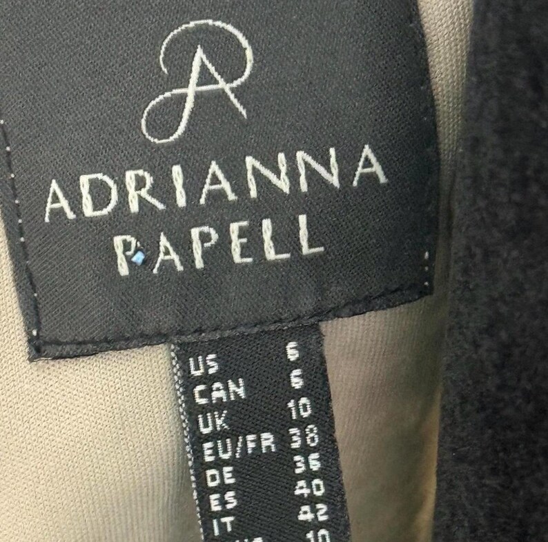 Pode incluir: Uma etiqueta preta com o nome da marca "ADRIANNA PAPELL" em letras brancas. Inclui convers&otilde;es de tamanhos para os tamanhos US, CAN, UK, EU/FR, DE, ES e IT. A etiqueta est&aacute; costurada numa pe&ccedil;a de vestu&aacute;rio.