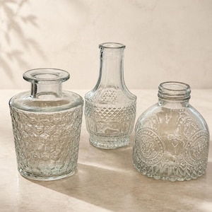 May include: Three clear glass bottles of varying shapes and sizes. The left bottle has a wide base with a hexagonal honeycomb pattern. The center bottle is tall with a diamond-textured body. The right bottle is rounded with embossed circular designs.