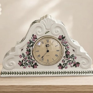 May include: A white ceramic mantel clock with a curved, stepped silhouette and embossed scroll details. The clock face features a cream background, black Roman numerals, and black hands. Pink floral and green leaf motifs decorate the front.