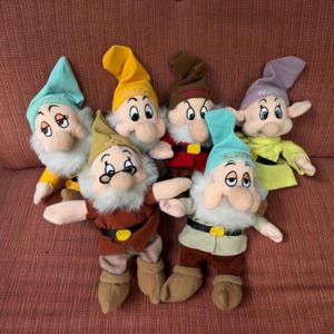 Puede incluir: Una colección de seis muñecos enanos de peluche, cada uno con un gorro puntiagudo bordado con un nombre. Los muñecos tienen barbas blancas, ropa de fieltro y cinturones negros con hebillas amarillas. Los personajes son Doc, Grumpy, Happy, Sleepy, Sneezy y Bashful.