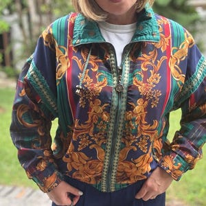 May include: A zip-up jacket featuring a vibrant pattern of gold ornate scrolls, cherubs, and vertical stripes in shades of navy blue, green, and red. The jacket has a high collar, a front zipper closure with a decorative pull, and long sleeves.