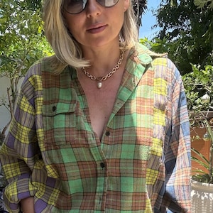 May include: A long-sleeve button-down shirt featuring a patchwork plaid pattern in shades of green, yellow, brown, and blue. The shirt has a collar, two chest pockets, and dark buttons. A gold-tone chain necklace with a single white pearl pendant.
