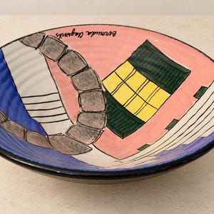 May include: A shallow ceramic bowl with a pink background featuring a hand-painted illustration of a stone archway, a yellow building with a green roof, and blue accents. The text Bermuda Clayworks is written in black script near the rim.
