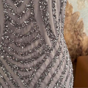 Pode incluir: Close-up de um vestido formal com uma base de tecido de malha cinza claro. O vestido &eacute; adornado com lantejoulas prateadas dispostas em um padr&atilde;o geom&eacute;trico. As lantejoulas s&atilde;o densamente compactadas, criando um efeito brilhante.