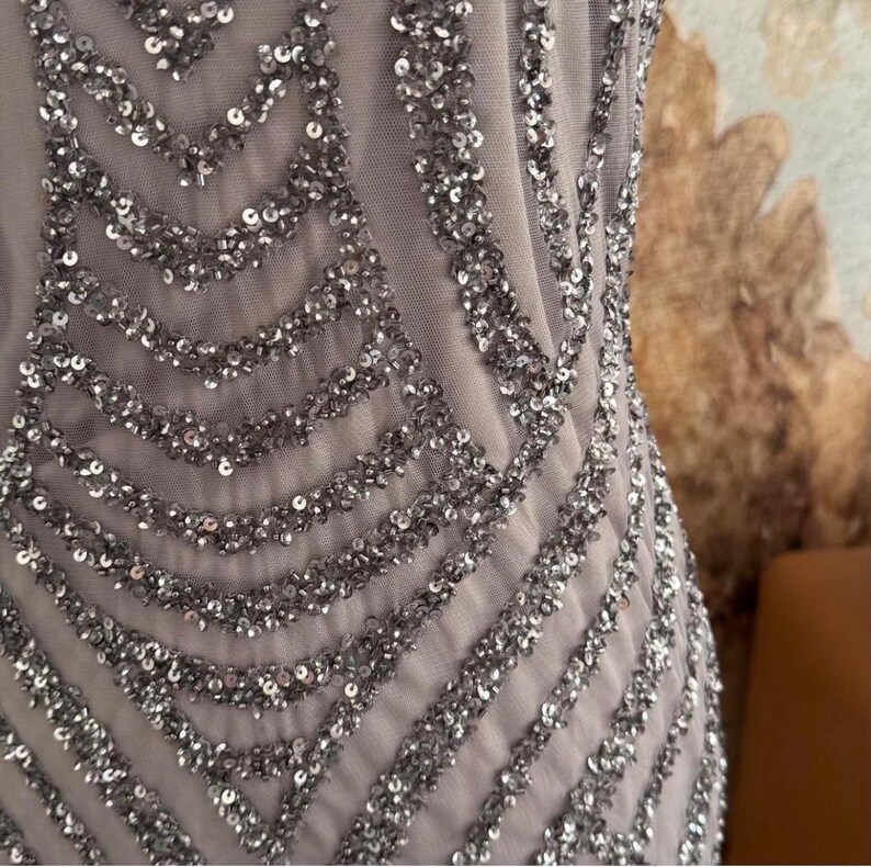 Pode incluir: Close-up de um vestido formal com uma base de tecido de malha cinza claro. O vestido &eacute; adornado com lantejoulas prateadas dispostas em um padr&atilde;o geom&eacute;trico. As lantejoulas s&atilde;o densamente compactadas, criando um efeito brilhante.
