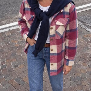 Puede incluir: Una camisa de cuadros en tonos burdeos, crema y azul marino, sobre una camiseta blanca lisa. Un suéter azul marino está colocado sobre los hombros y anudado en la parte delantera. El conjunto incluye vaqueros azules y un cinturón de cuero marrón.