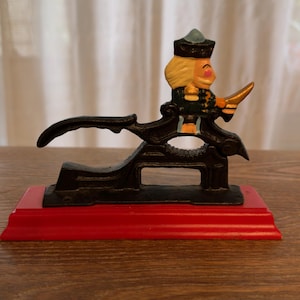 Op de afbeelding: Een zwarte gietijzeren notenkraker met een geschilderde soldatenfiguur met een beige gezicht, blond haar en een donkergroen uniform. De figuur is gemonteerd op een zwart metalen hefboommechanisme, bevestigd op een rode houten basis.