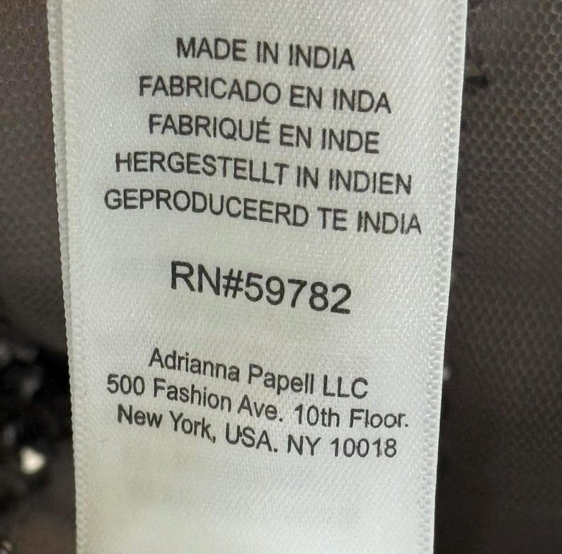 Pode incluir: Close-up de uma etiqueta de tecido branco com texto em v&aacute;rios idiomas: Made in India, Fabricado en Inda, Fabriqu&eacute; en Inde, Hergestellt in Indien, Geproduceerd te India. A etiqueta tamb&eacute;m inclui o texto RN#59782 e Adrianna Papell LLC, 500 Fashion Ave., New York, USA, NY 10018.