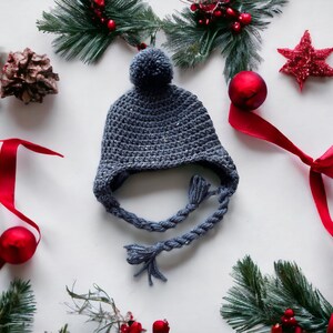May include: A gray crocheted hat with a pompom on top and a braided cord. The hat is surrounded by red ornaments, pine branches, and red ribbon.