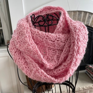 Peut inclure: Une écharpe en crochet infinie rose. L'écharpe est réalisée avec un point texturé et est présentée sur un mannequin noir.