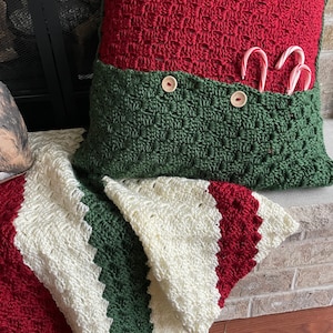 May include: A green and red crocheted pillow with a pocket for candy canes. The pillow is sitting on a white and green striped crocheted blanket.