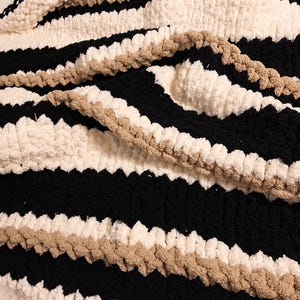 May include: A chunky knit blanket with alternating stripes of black, cream, and tan. The soft, textured blanket is folded, showcasing the thick yarn and color pattern. This cozy throw is perfect for a bed or sofa.