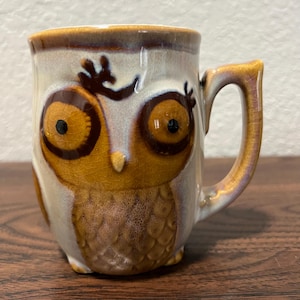 May include: A ceramic mug shaped like an owl with a glossy, crackled glaze finish. The mug features a cream-colored body with brown accents around the large, circular eyes, a small pointed beak, and textured feather patterns on the front surface.