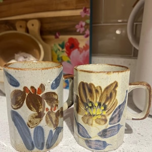 May include: Two ceramic mugs with floral designs. One mug features brown and orange abstract flowers with blue leaves. The other mug has a large brown flower with yellow center and blue leaves. Both mugs have a beige background and brown handles.