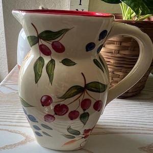 Op de afbeelding: Crèmekleurige keramische kan met een rode rand en een gebogen handvat. De kan is versierd met groene bladeren en trossen rode en blauwe bessen. Een decoratief item met de letters Z en I staat op de achtergrond.