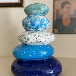 May include: A stacked ceramic sculpture with a pebble-like design. The sculpture features a gradient of colors, starting with a deep blue base, transitioning to lighter blues, and topped with a turquoise element. The surfaces have speckled patterns.