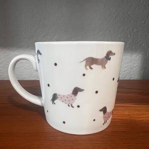 Könnte beinhalten: Ein weißer Keramikbecher mit einem wiederkehrenden Muster aus braunen Dackeln. Einige Hunde tragen kleine rosa Pullover mit Punkten, während andere dunkle Halsbänder haben. Der Becher ist mit verstreuten kleinen schwarzen Punkten verziert.
