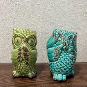 May include: Two ceramic owl figurines with a textured, scale-like pattern. The left owl is lime green and covers its mouth with its wings. The right owl is turquoise and covers its eyes with its wings. Both feature dark brown distressed accents.