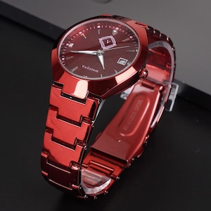 Könnte beinhalten: Eine rote Armbanduhr mit passendem Armband liegt auf einer schwarzen Oberfläche neben einem Laptop und einem Smartphone. Das Zifferblatt der Uhr ist dunkelrot mit silbernen Zeigern und Markierungen.