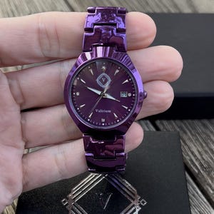 May include: A purple watch with a matching band. The watch face has a dark purple background with silver hands and markers. The watch face also has the text "Valirium" and a date display. The watch is held in a hand.