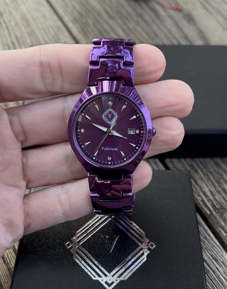 May include: A purple watch with a matching band. The watch face has a dark purple background with silver hands and markers. The watch face also has the text "Valirium" and a date display. The watch is held in a hand.