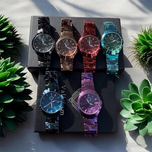 May include: Six colorful watches with matching bands are displayed on a black box. The watches have round faces and are in shades of black, bronze, red, teal, blue, and purple. The watches are surrounded by green plants.