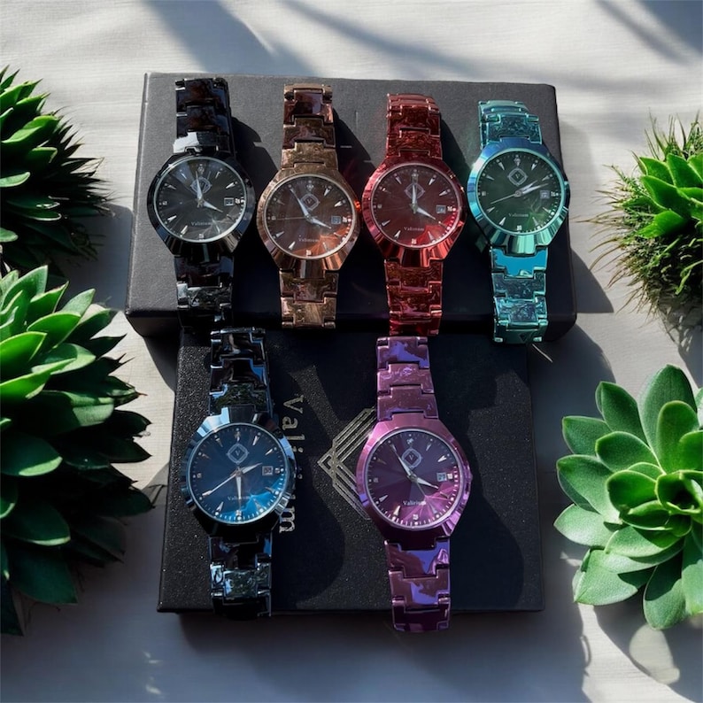 May include: Six colorful watches with matching bands are displayed on a black box. The watches have round faces and are in shades of black, bronze, red, teal, blue, and purple. The watches are surrounded by green plants.