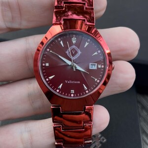 May include: A red wristwatch with a matching band. The watch face is a deep red with diamond-like markers and silver hands. The watch face has the text "Valirium" and a date display. The watch is held in a hand.