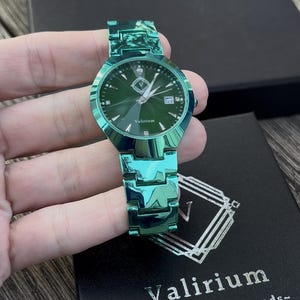 May include: A green metallic watch with a matching band. The watch face is dark green with silver hands and markers. The watch has the brand name "Valirium" on the face. The watch is on a black box with the brand name and tagline.
