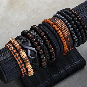 Może przedstawiać: Kolekcja bransoletek w różnych stylach, w tym koraliki, pleciona skóra i skórzane wzory owijane. Bransoletki charakteryzują się mieszanką kolorów, w tym brązu, czerni i naturalnych odcieni drewna. Symbol nieskończoności jest również dołączony.