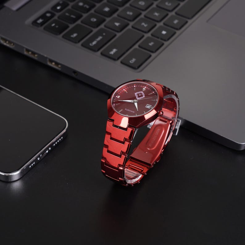 May include: A red wristwatch with a matching band sits on a black surface next to a laptop and a smartphone. The watch has a dark red face with silver hands and markers. The band is made of interlocking metal links.
