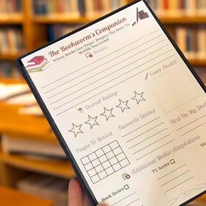 May include: A black and white book journal titled "The Bookworm's Companion." The journal includes sections for overall ratings, favourite quotes, and pages to revisit. The cover features a book and a pencil illustration. The background is a blurred library.