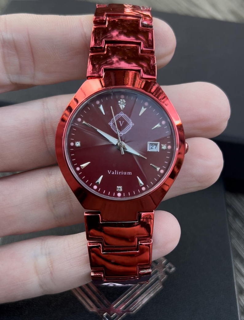 May include: A red wristwatch with a matching band. The watch face is a deep red with diamond-like markers and silver hands. The watch face has the text "Valirium" and a date display. The watch is held in a hand.