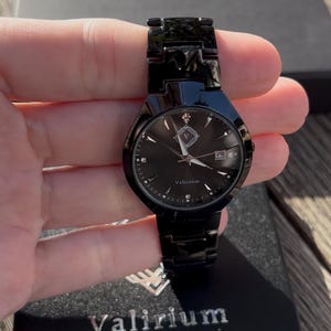May include: A black wristwatch with a matching band. The watch face is black with silver hands and markers, and a date window at the 3 o'clock position. The watch is held in a hand, and the brand name "Valirium" is visible on the watch face and box.