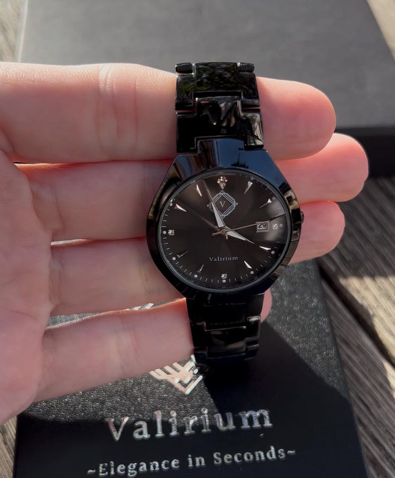 May include: A black wristwatch with a matching band. The watch face is black with silver hands and markers, and a date window at the 3 o'clock position. The watch is held in a hand, and the brand name "Valirium" is visible on the watch face and box.