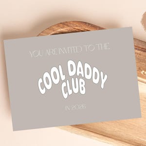 May include: A gray card with the text "YOU ARE INVITED TO THE COOL DADDY CLUB IN 2028" in white. The card is on a wooden tray with dried flowers and a round sticker that says "DIGITALES PRODUKT".