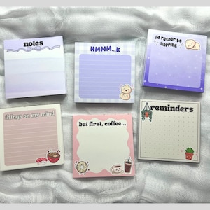 May include: A collection of six square sticky note pads with various designs and text. The pads feature pastel colors, illustrations of food and cute characters, and phrases like "notes," "HMMM...K," and "I'd rather be napping."