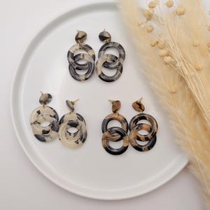 Pode incluir: Quatro pares de brincos pendurados com um design marmorizado em tons de preto, castanho e creme. Os brincos apresentam formas circulares interligadas e são exibidos em um prato branco. Os brincos têm pinos dourados.