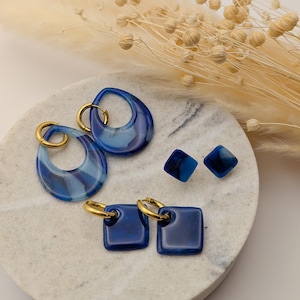 Può includere: Una collezione di orecchini in resina blu con cerchi dorati. Gli orecchini includono modelli a goccia, quadrati e a bottone, che mostrano sfumature di blu e bianco. I gioielli sono esposti su una superficie marmorizzata con accenti floreali secchi.