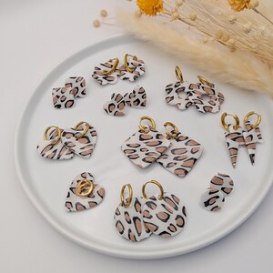 Peut inclure: Une collection de boucles d'oreilles faites à la main avec un motif léopard blanc, noir et beige. Les boucles d'oreilles sont de différentes formes, notamment des cœurs, des carrés et des cercles, et comportent des cerceaux dorés. Les boucles d'oreilles sont présentées sur une assiette blanche.