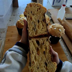 May include: A cross-section of a golden-brown panettone, revealing a light, airy interior with dried fruit inclusions. The bread is held in front of other baked goods, including a muffin-shaped cake with white sugar crystals.