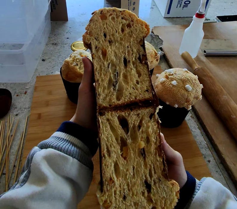 May include: A cross-section of a golden-brown panettone, revealing a light, airy interior with dried fruit inclusions. The bread is held in front of other baked goods, including a muffin-shaped cake with white sugar crystals.