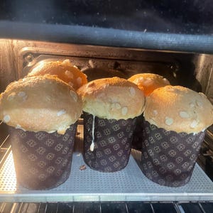 May include: Several freshly baked, golden-brown panettone cakes with almond toppings are in an oven. The cakes are in decorative paper molds with a dark brown floral pattern. The oven light illuminates the baked goods.