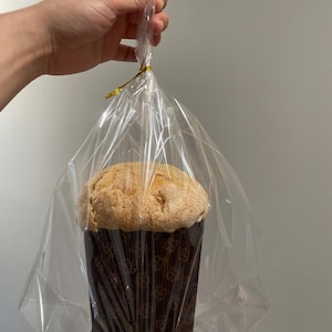 Può includere: Un prodotto da forno grande e rotondo, forse una torta o del pane, in una tazza di carta marrone con un motivo floreale. Il prodotto da forno è avvolto in cellophane trasparente e legato con un nastro dorato. L'immagine è di una mano che tiene il prodotto da forno confezionato.