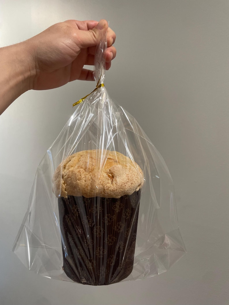 May include: A large, round baked good, possibly a cake or bread, in a brown paper cup with a floral design. The baked good is wrapped in clear cellophane and tied with a gold ribbon. The image is of a hand holding the wrapped baked good.